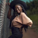 Stylish woman with braids and hat poses confidently near a fence in Ghana.
